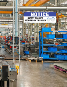 Overhead safety sign titled "Safety Glasses Required In This Area" printed on 4mm corrugated plastic with nickel grommets and ceiling grid clips.