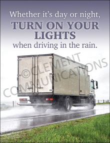 A commercial box truck driving on a wet road during a rainstorm with water splashing from the tires, beneath bold text reading: "Whether it's day or night, TURN ON YOUR LIGHTS when driving in the rain."